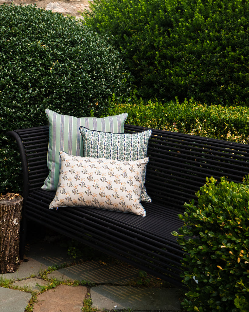 Three decorative outdoor throw pillows in green, beige, and patterned designs resting on a black metal garden bench surrounded by lush green hedges.