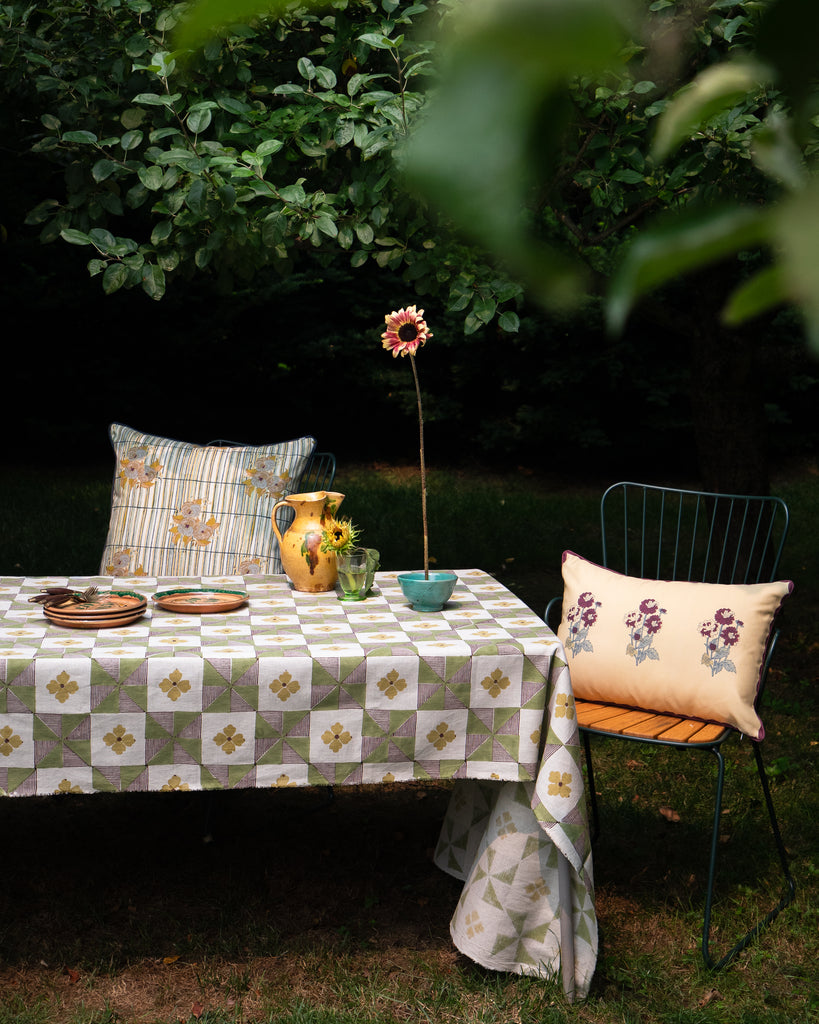 Outdoor garden table setting with a checkered tablecloth, embroidered throw pillows, a yellow ceramic pitcher, and a green wire chair on grass.