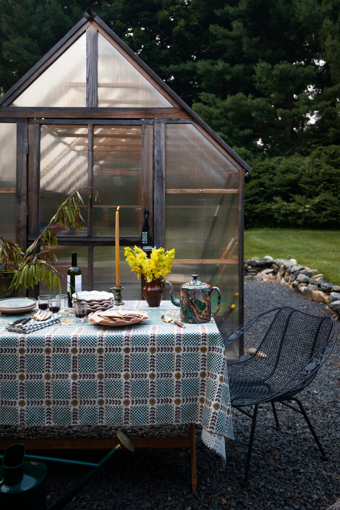 An outdoor dining setup with a patterned tablecloth, plates, candles, and yellow flowers in front of a greenhouse, with a black wire chair beside it and trees in the background.