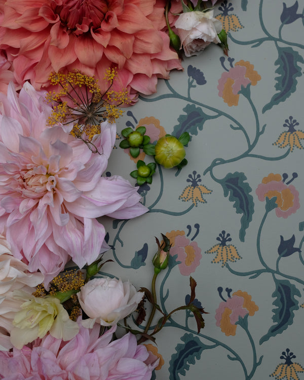 A styled flat-lay of fresh flowers, including pink and coral dahlias, roses, and green seed pods, arranged on top of a light gray wallpaper with a vintage floral vine pattern.