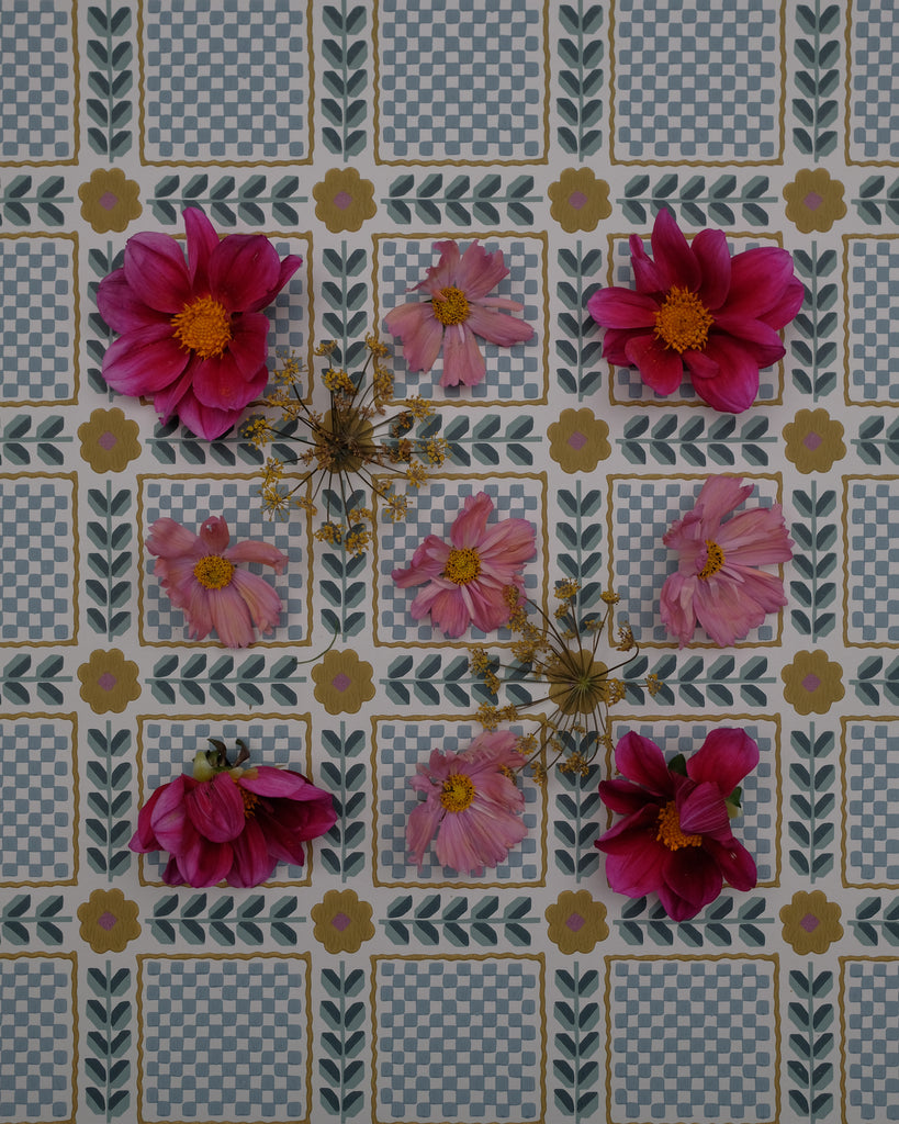 A close-up of a patterned fabric or wallpaper featuring a grid of blue-and-white checkered squares, olive-green leafy borders, and scattered pink and magenta flowers with yellow centers.