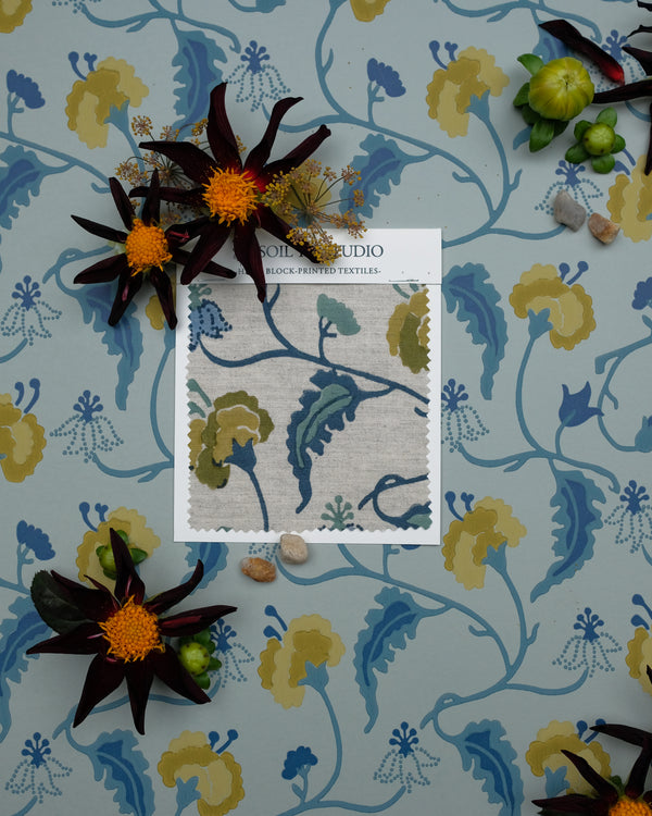 A fabric swatch card from Wool Studio is displayed on a matching floral wallpaper background, surrounded by decorative dark flowers and small stones. The swatch features a botanical pattern in muted blue, olive green, and beige tones.