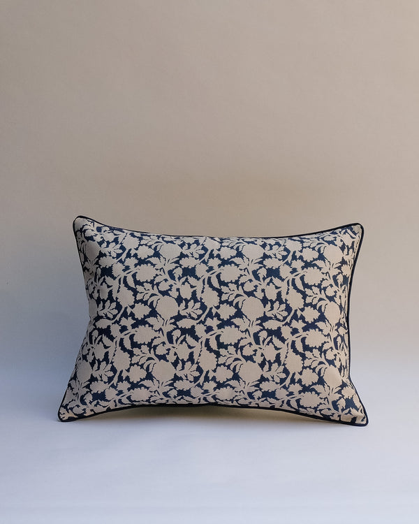A rectangular decorative throw pillow with a navy blue and beige floral damask pattern, finished with dark piping, set against a neutral beige background.
