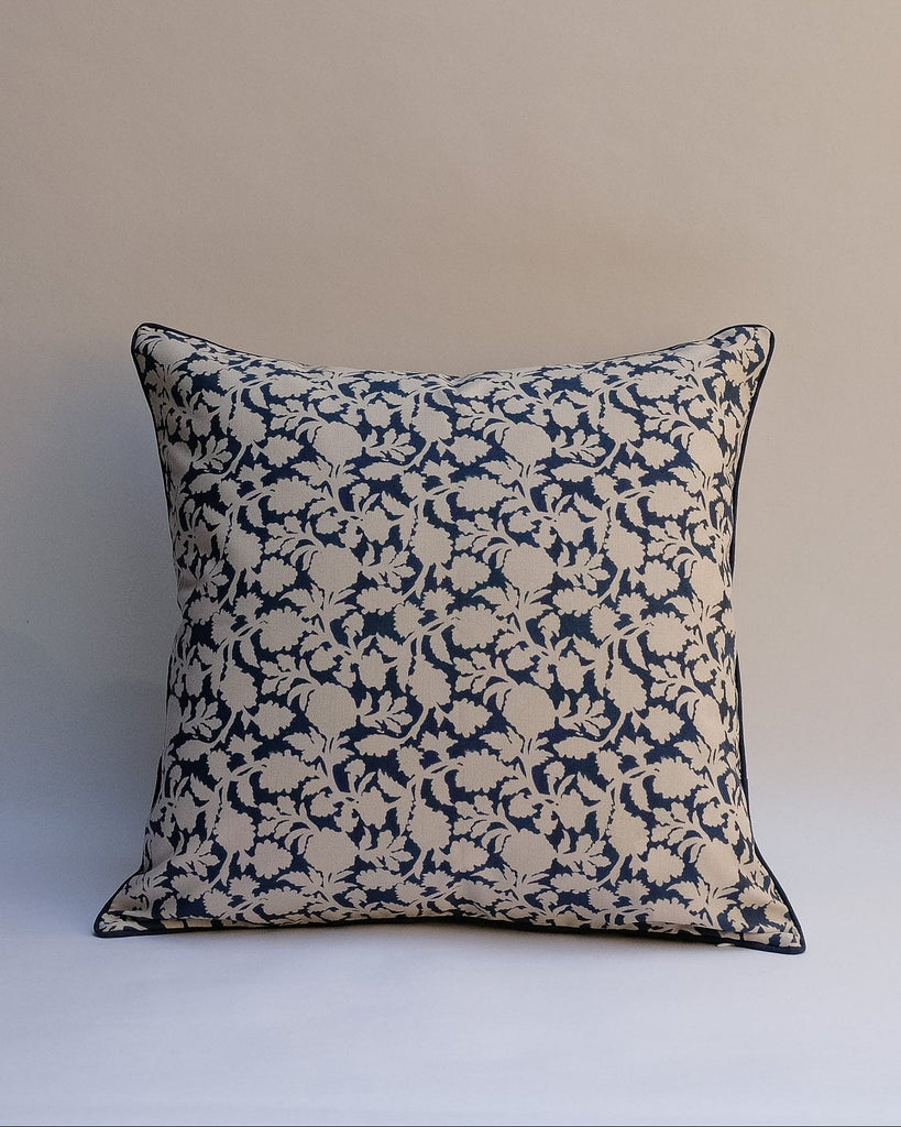 A square throw pillow with a navy blue and beige floral damask pattern, finished with a thin dark piped edge, set against a neutral beige background.