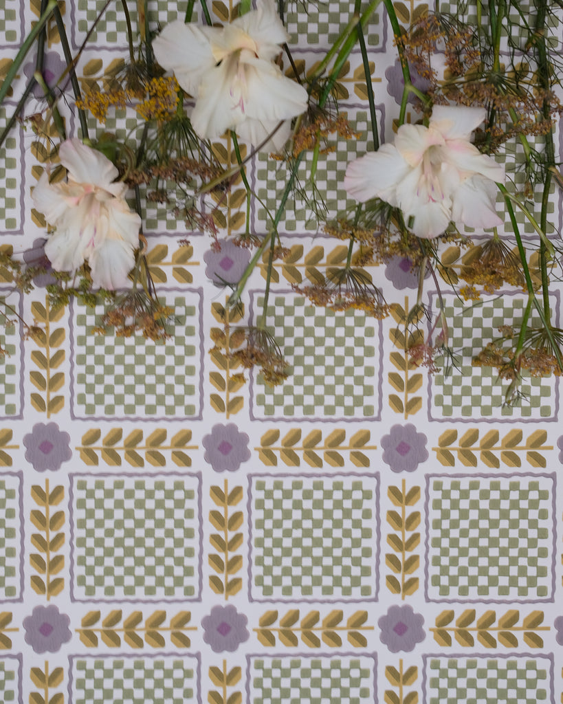 A close-up of a patterned fabric or wallpaper featuring a repeating grid of green and white checkered squares bordered by yellow leaf motifs and small lavender flowers, with pale pink flowers and green stems laid across the top.