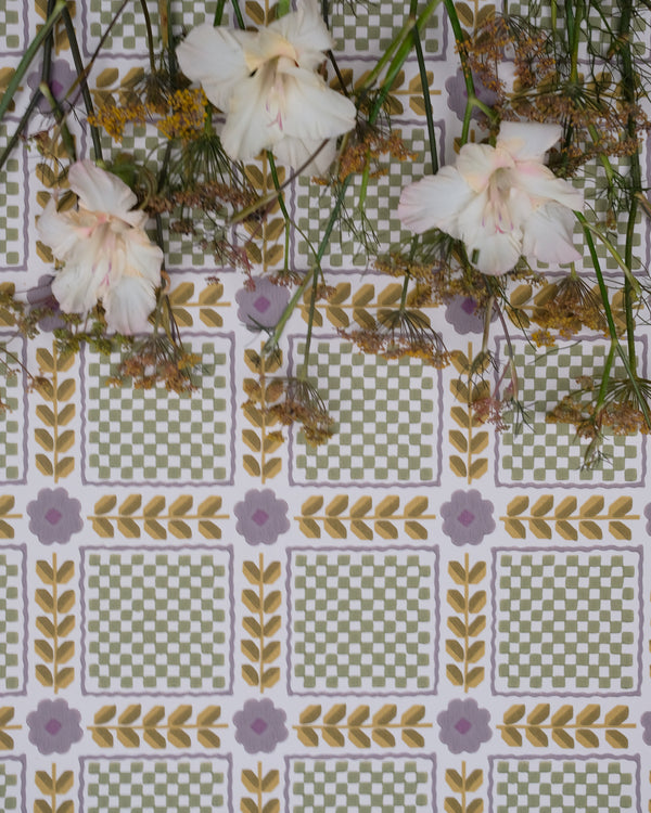 A close-up of a patterned fabric or wallpaper featuring a repeating grid of green and white checkered squares bordered by yellow leaf motifs and small lavender flowers, with pale pink flowers and green stems laid across the top.