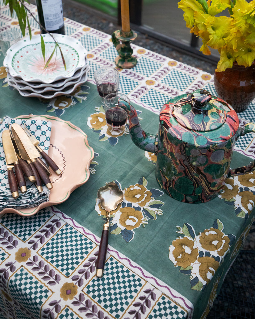 A styled table setting featuring a green and gold floral patterned tablecloth, a marbled green teapot, stacked white plates, a pink ceramic plate with knives, and yellow flowers in a vase.