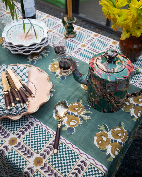 A styled table setting featuring a green and gold floral patterned tablecloth, a marbled green teapot, stacked white plates, a pink ceramic plate with knives, and yellow flowers in a vase.