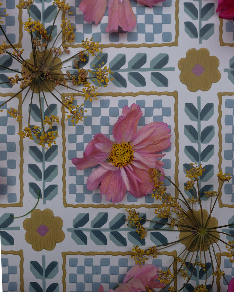 A close-up of a decorative surface featuring a repeating geometric tile pattern in muted blue, beige, and olive tones, overlaid with pressed pink flowers and small yellow blossoms.