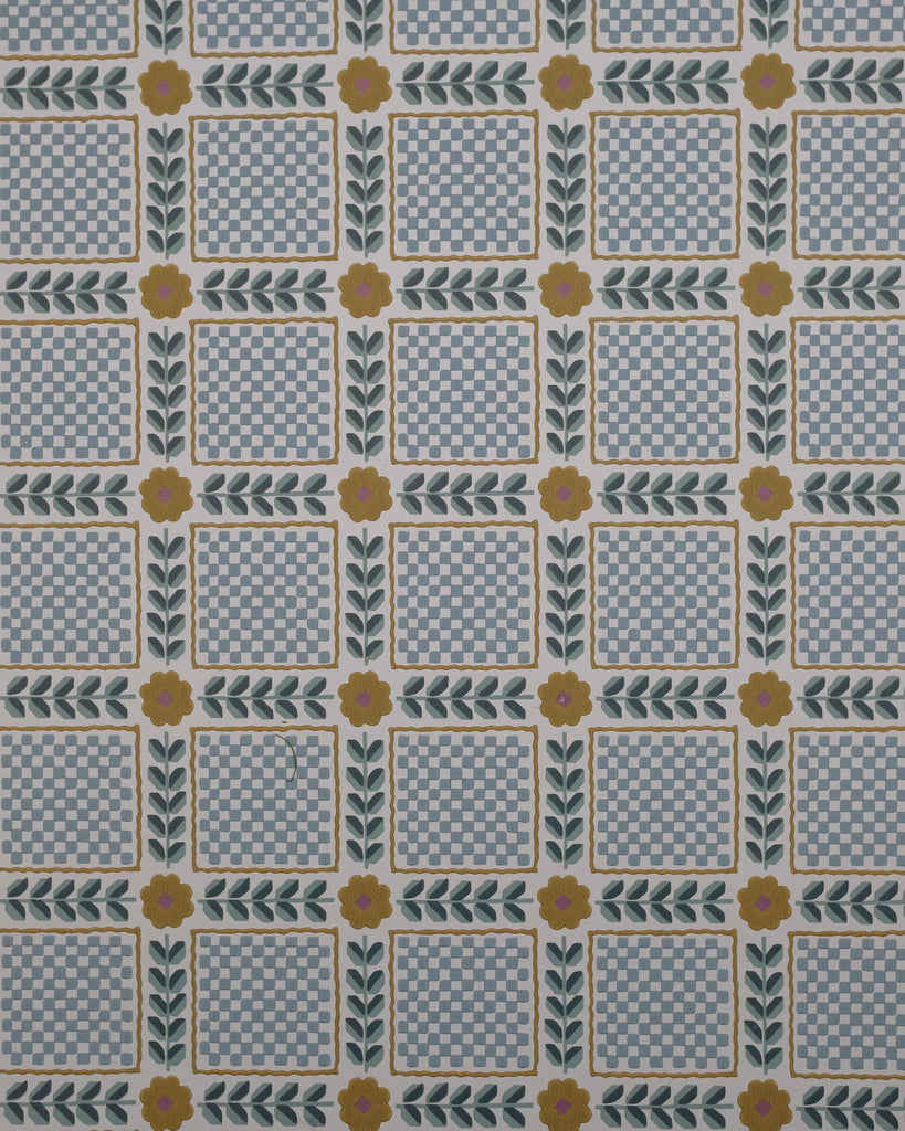Mumtaz wallpaper finds beauty in order and repetition, with echoes of vintage patchwork and old hand-worked textiles. Tiny checks sit within leafy borders, punctuated by simple flowers, creating a rhythm that feels both nostalgic and playful. There is a quiet charm to its geometry—the kind of pattern that reveals more of itself the longer you live with it.