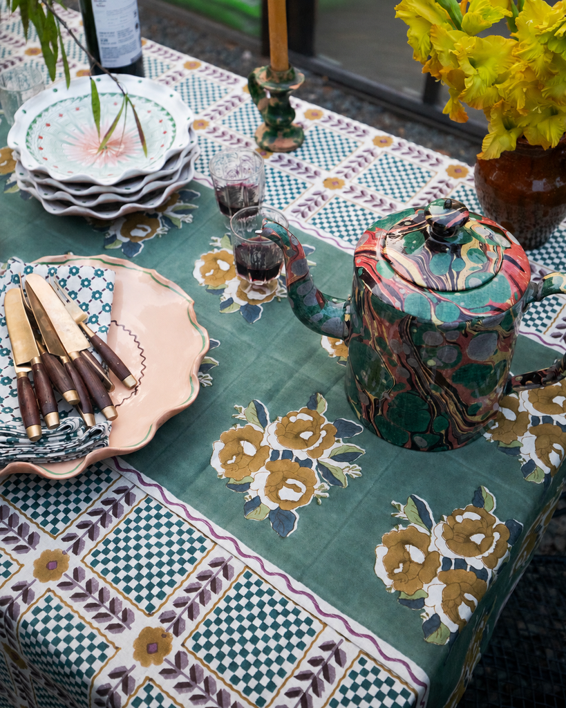 A styled table setting featuring a green tablecloth with a floral and geometric patchwork pattern, a matching patterned teapot, stacked white plates, a pink platter with wooden-handled knives, and yellow flowers in a vase.