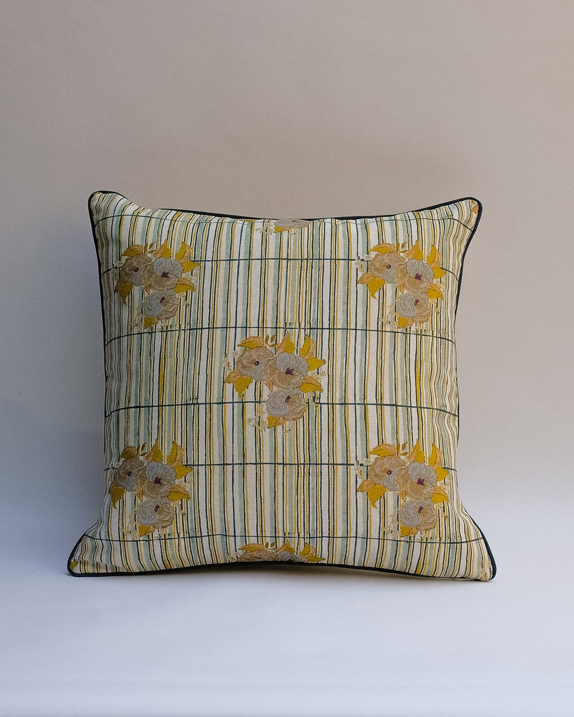 A square decorative outdoor pillow with a retro floral pattern in muted shades of pink, purple, olive green, and beige, finished with dark brown piping.