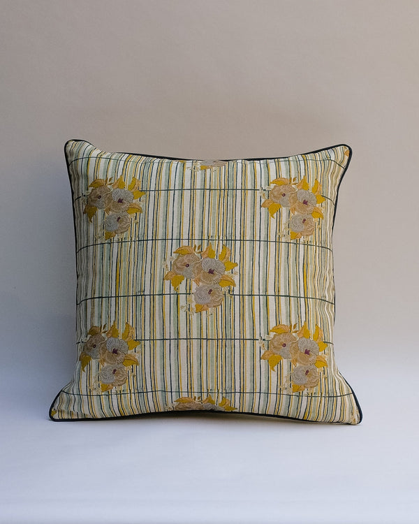 A square decorative outdoor pillow with a retro floral pattern in muted shades of pink, purple, olive green, and beige, finished with dark brown piping.