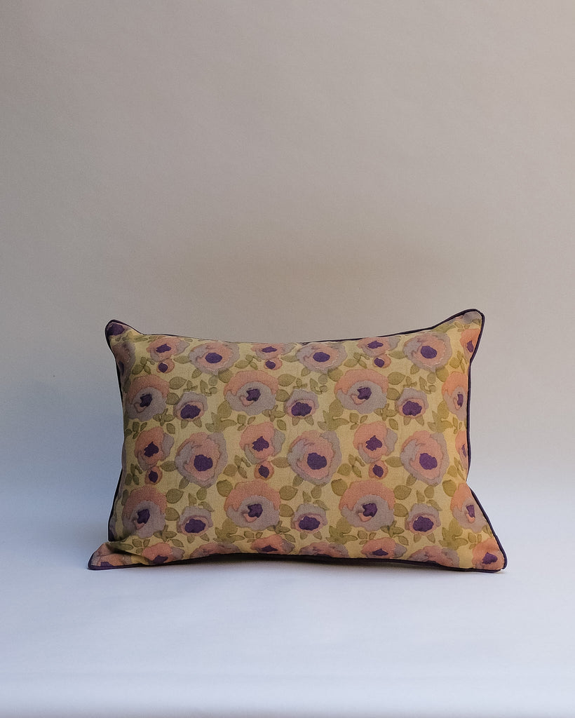 A decorative outdoor pillow with a retro floral pattern in muted shades of pink, purple, olive green, and beige, finished with dark brown piping.
