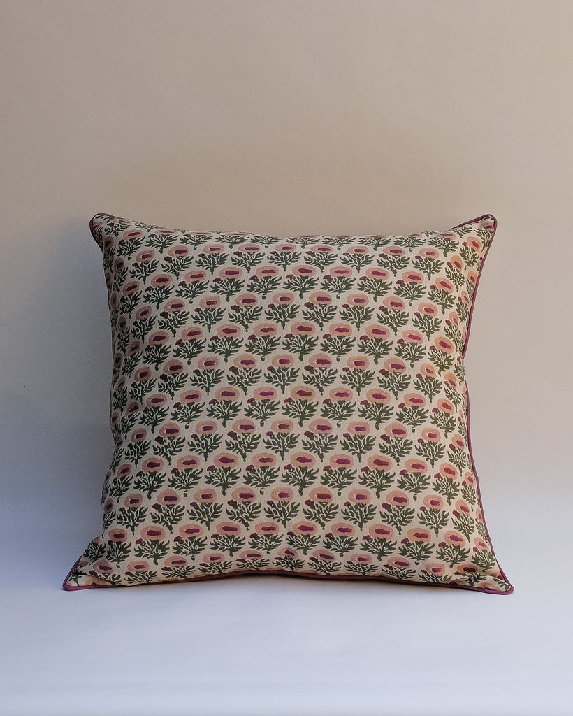 A square throw pillow featuring a repeating block-print style floral pattern in green and pink on a cream background, with a thin dark pink piped edge, set against a plain light beige wall.