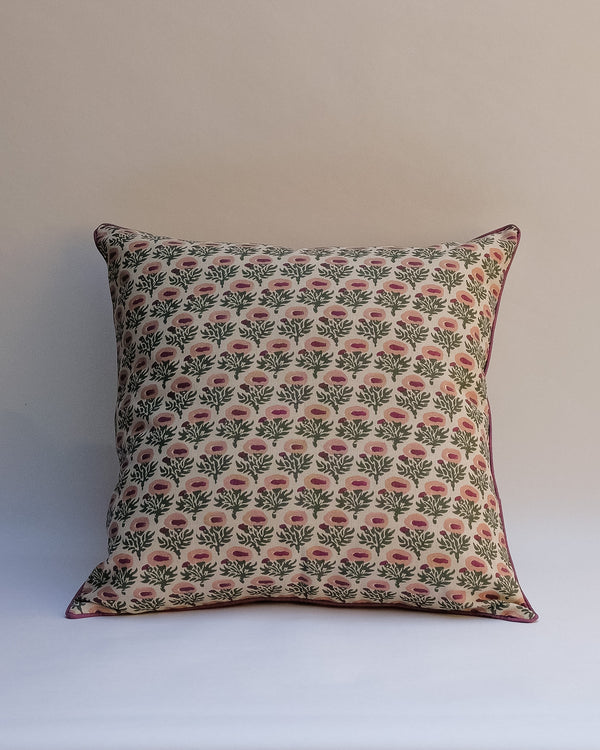 A square throw pillow featuring a repeating block-print style floral pattern in green and pink on a cream background, with a thin dark pink piped edge, set against a plain light beige wall.