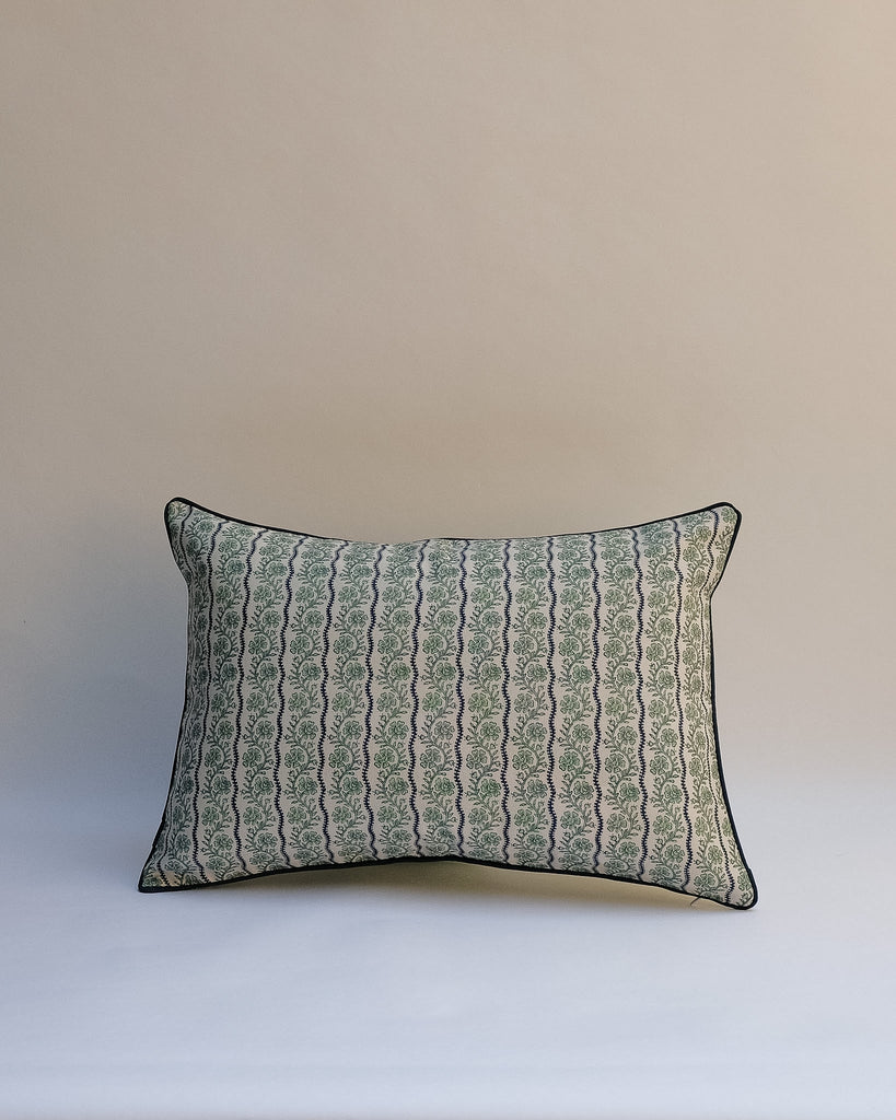A rectangular decorative throw pillow with a vertical wavy pattern in muted sage green, gray, and beige, finished with a thin black piped edge, set against a plain beige background.