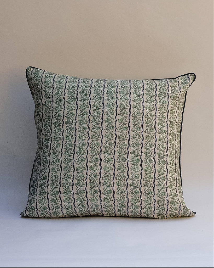 A square decorative throw pillow with a vertical wavy pattern in sage green and beige, finished with a thin black piped edge, set against a plain beige background.