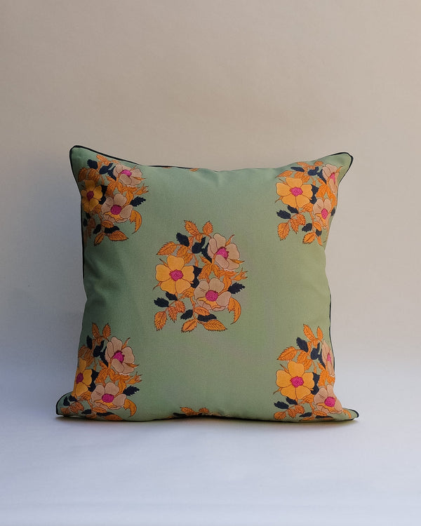 A square decorative outdoor pillow with a retro floral pattern in muted shades of pink, purple, olive green, and beige, finished with dark brown piping.