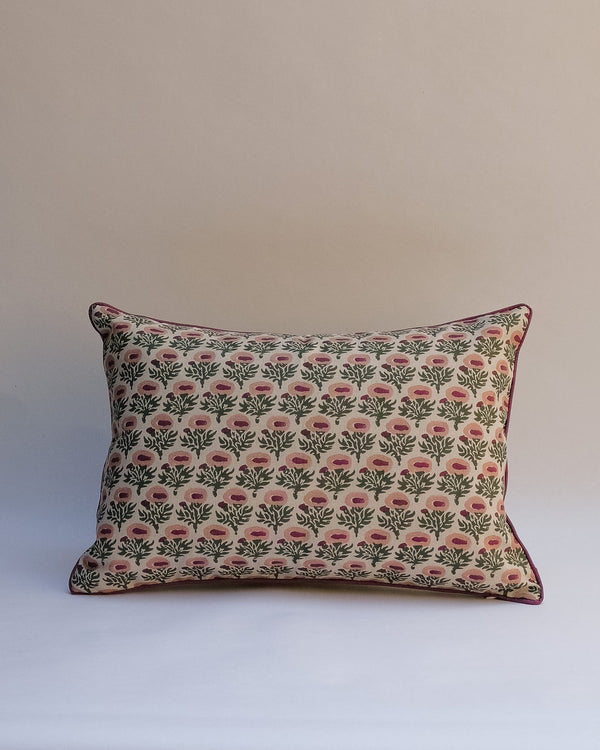A rectangular decorative throw pillow with a repeating floral block-print pattern in pink and green on a cream background, finished with a dark pink piped edge, set against a plain beige studio backdrop.